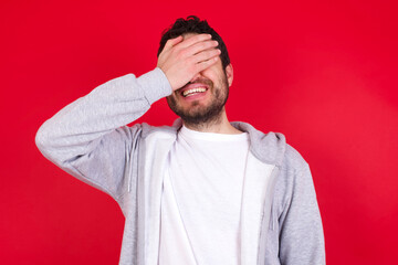 Happy young handsome caucasian man in sports clothes against red wall closing eyes with hand going to see surprise prepared by friend standing and smiling in anticipation for something wonderful.