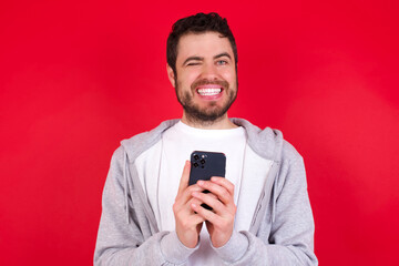 Pleased young handsome caucasian man in sports clothes against red wall using self phone and looking and winking at the camera. Flirt and coquettish concept.
