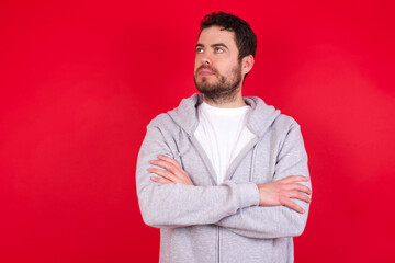 Charming thoughtful young handsome caucasian man in sports clothes against red wall stands with arms folded concentrated somewhere with pensive expression thinks what to do