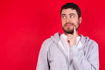 Thoughtful young handsome caucasian man in sports clothes against red wall holds chin and looks away pensively makes up great plan