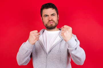 Displeased annoyed young handsome caucasian man in sports clothes against red clenches fists, gestures pissed, ready to revenge, looks with aggression at camera stands full of hate, being pressured