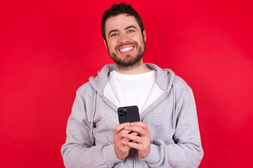 young handsome caucasian man in sports clothes against red wall Mock up copy space. Using mobile phone, typing sms message