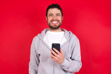 young handsome caucasian man in sports clothes against red wall holds mobile phone in hands and rejoices positive news, uses modern cellular
