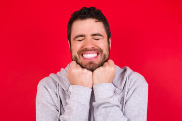 Cheerful young handsome caucasian man in sports clothes against red background has shy satisfied expression, smiles broadly, shows white teeth, People emotions