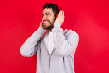 young handsome caucasian man in sports clothes against red background wears stereo headphones listens music concentrated aside. People hobby lifestyle concept