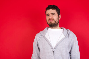Displeased young handsome caucasian man in sports clothes against red background frowns face feels unhappy has some problems. Negative emotions and feelings concept