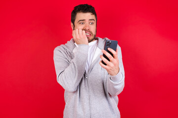 Portrait of pretty frightened young handsome caucasian man in sports clothes against red background chatting biting nails after reading some scary news on her smartphone.