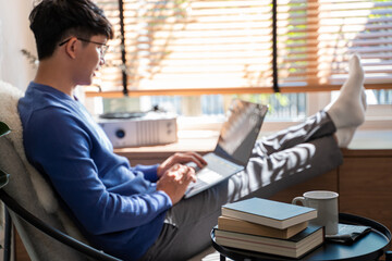 Young asian male tech user relaxing  holding laptop computer and looking at the screen in living room, Remote Job or work from home concept.