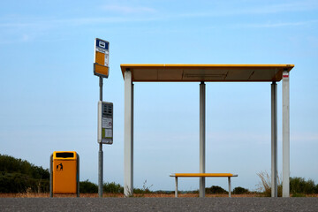 Bus stop in Holland, pillars support yellow roof, small bench underneath, gray street in the...