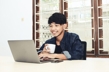 Asian man sits in front of a laptop drinking coffee