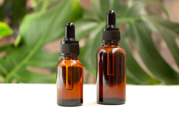 Two amber glass bottles stand on a white table against a background of monstera leaves. Natural organic eco-cosmetics. Beauty salon branding mockup. Aromatherapy.