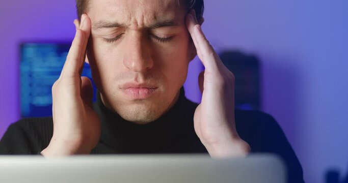 Close-up stressed young unhealthy man touching temples, suffering from headache migraine pain while working at laptop computer. Concept of overworking