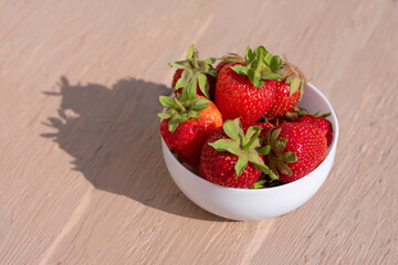 White bowl full of bright red ripe strawberries on hard sunlight and long shadows on wooden background.