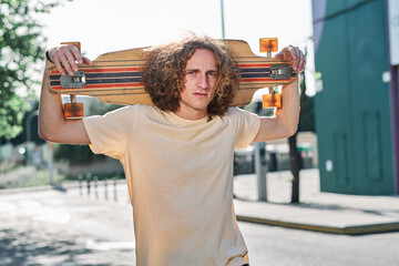 young man with curly hair looking at camera with his longboard or skeateboard on his shoulders in...