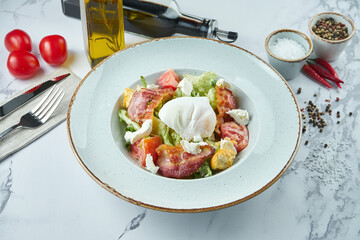 Appetizing caesar or lardon salad with croutons, bacon, feta cheese, tomatoes and poached egg in a blue plate on a marble background.