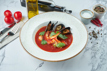 Appetizing Spanish tomato soup with seafood. Red soup with tiger prawns, mussels and basil in a white plate on a marble background.