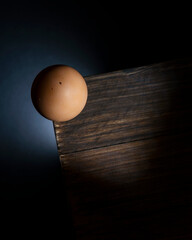 Top view of a chicken egg precariously balanced on the corner of a wooden table as it is about to fall to the ground