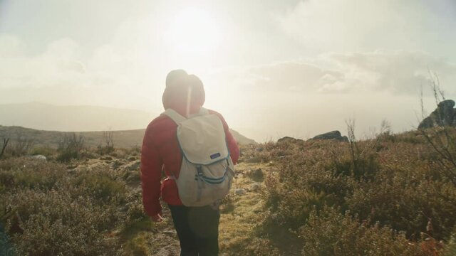 Cinematic and inspiring shot of man in red puffy jacket with canvas backpack walk on beautiful epic trail high up in mountains. Inspirational travel wanderlust concept. Reaching summit at sunrise 