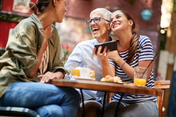 A group of female friends of different generations are laughing at smartphone content they watching while they have a drink in the bar. Leisure, bar, friendship, outdoor