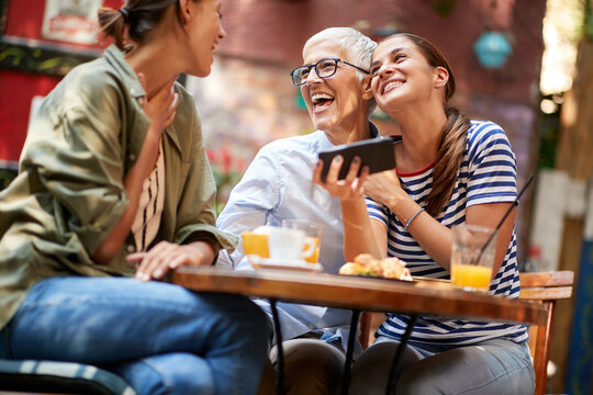 A Group Of Female Friends Of Different Generations Are Laughing At Smartphone Content They Watching While They Have A Drink In The Bar. Leisure, Bar, Friendship, Outdoor