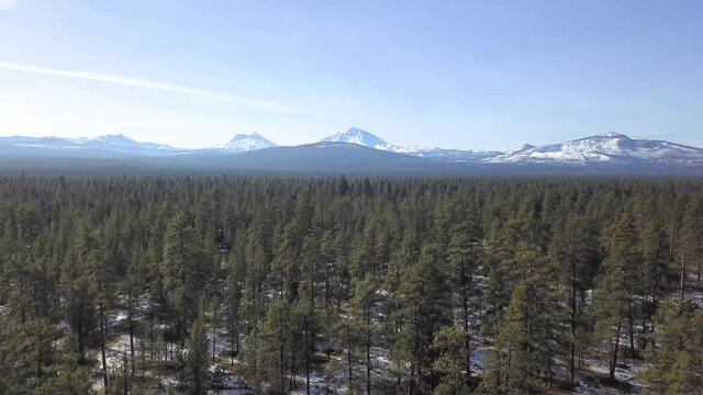Aerial flying backwards and ascending above Douglas Fir forest in central Oregon and the Three Sisters mountains covered in snow in the distance.