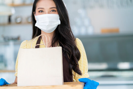 Attractive Asian Female Cafe Worker Wears Face Mask And Gloves Giving Takeaway Food Bag To Customer.waitress Holding Takeout Order Standing In Coffee Shop Front Door Entrance New Normal Lifestyle