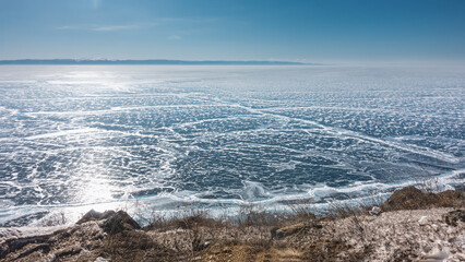 The smooth surface of the frozen lake is lined with cracks and covered with snow patterns. Sun glare on ice. Dry grass on the shore. Winter sunny day. Blue sky. Baikal