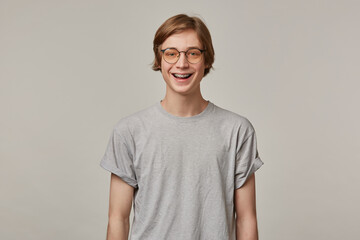 Happy looking man, handsome guy with blond hair. Wearing grey t-shirt, glasses and has braces. People and emotion concept. Watching and broadly smiling at the camera isolated over grey background