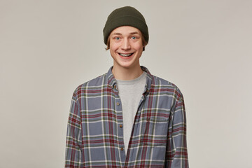 Cheerful guy, happy looking man with blond hair. Wearing checkered shirt and beanie. Has braces. People and emotion concept. Watching positive at the camera isolated over grey background