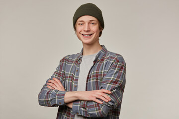 Cool looking male, handsome guy with blond hair. Wearing checkered shirt and beanie. Has braces. Holds arms crossed. People and emotion concept. Watching at the camera isolated over grey background