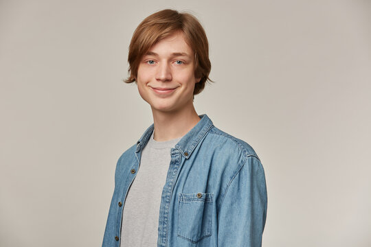 Positive teenage guy, happy looking man with blond hair. Wearing blue denim shirt. People and emotion concept. Watching and smiling at the camera isolated over grey background