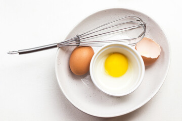 Egg yolk in small bowl. Chicken egg, eggshells and whisk in large white bowl.