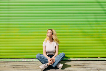 Young blonde woman wearing a white top and blue jeans, sitting on a wooden floor on a vivid green wall background.