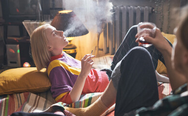 Young woman lying on bed and smoking a cigarette with her boyfriend sitting on the bed and drinking wine