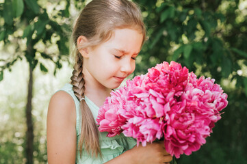 Fototapeta premium portrait of a happy cute little caucasian seven year old kid girl, holds in hands and smell and enjoying a bouquet of pink peony flowers in full bloom on the background of nature