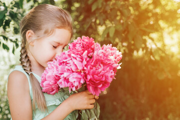 Fototapeta premium portrait of a happy cute little caucasian seven year old kid girl, holds in hands and smell and enjoying a bouquet of pink peony flowers in full bloom on the background of nature. flare