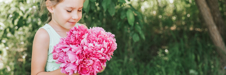 Fototapeta premium portrait of a happy cute little caucasian seven year old kid girl, holds in hands and smell and enjoying a bouquet of pink peony flowers in full bloom on the background of nature. banner