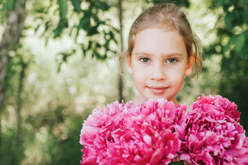 Fototapeta premium portrait of a happy cute little caucasian seven year old kid girl, holds in hands a bouquet of pink peony flowers in full bloom on the green background of nature
