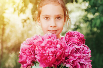 Naklejka premium portrait of a happy cute little caucasian seven year old kid girl, holds in hands a bouquet of pink peony flowers in full bloom on the green background of nature. flare