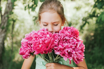 Fototapeta premium portrait of a happy cute little caucasian seven year old kid girl, holds in hands and smell and enjoying a bouquet of pink peony flowers in full bloom on the background of nature