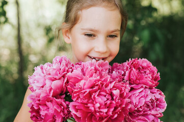 Fototapeta premium portrait of a happy cute little caucasian seven year old kid girl, holds in hands and smell and enjoying a bouquet of pink peony flowers in full bloom on the background of nature