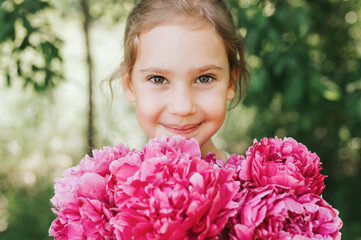 Fototapeta premium portrait of a happy cute little caucasian seven year old kid girl, holds in hands a bouquet of pink peony flowers in full bloom on the green background of nature