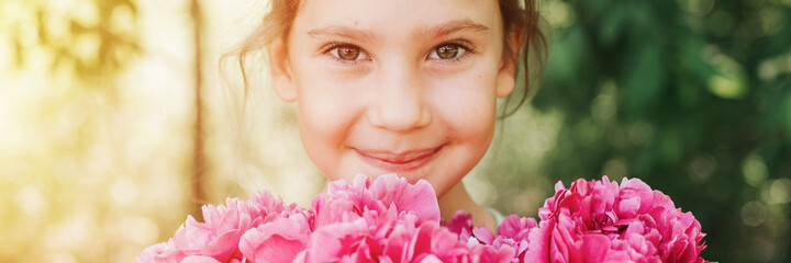 Fototapeta premium portrait of a happy cute little caucasian seven year old kid girl, holds in hands a bouquet of pink peony flowers in full bloom on the green background of nature. banner. flare