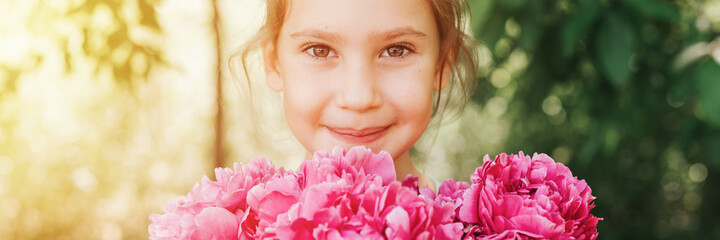 Fototapeta premium portrait of a happy cute little caucasian seven year old kid girl, holds in hands a bouquet of pink peony flowers in full bloom on the green background of nature. banner. flare