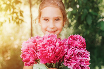 Fototapeta premium portrait of a happy cute little caucasian seven year old kid girl, holds in hands a bouquet of pink peony flowers in full bloom on the green background of nature
