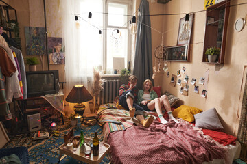 Young woman lying on the bed and reading a book with her boyfriend sitting near by her and smoking at home