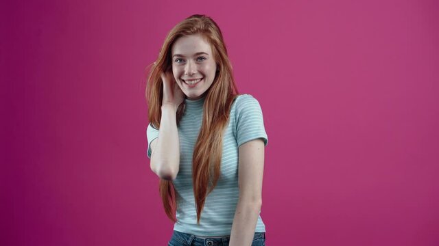 The red-haired teenager listens to the new rumor that surprises her and makes her smile, in a casual blue T-shirt, isolated on a pink background. The concept of people's lifestyle