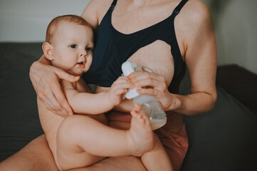 Young mother with newborn cute infant naked baby boy, holding him on arms, using breast pump hugging and breastfeeds with breast milk. Healthy child, concept of hospital and happy motherhood. Nursery