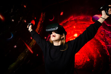 The girl cheerfully and emotionally rejoices at the victory in a video game wearing virtual reality glasses, holding gamepads in her hands. Red game background.