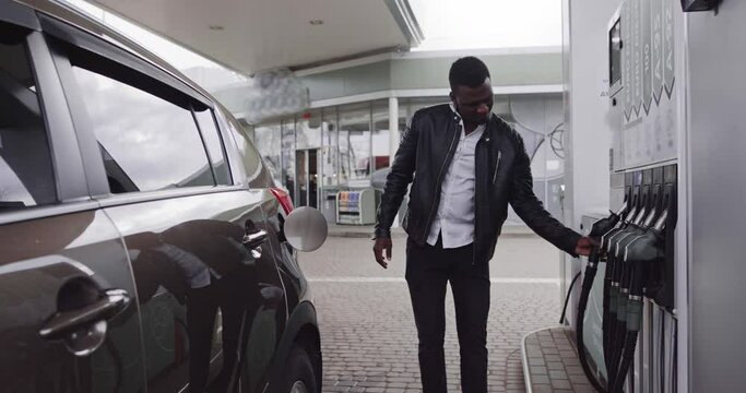 Young handsome African bearded guy in stylish casual outfit refueling his luxury car and looking away while standing on self service petrol station. Translational: ENERGY OF YOUR MOVEMENT